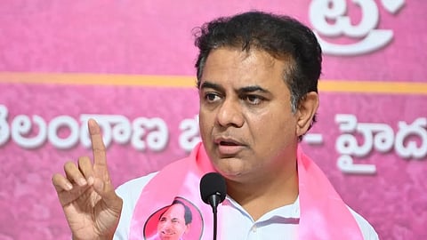 A close-up shot of Indian politician K. T. Rama Rao (KTR) speaking at a podium. He is wearing a light-colored button-down shirt and a bright pink Bharat Rashtra Samithi (BRS) party scarf draped over his shoulders. The scarf features the party's name in multiple languages and a portrait of his father, K. Chandrashekar Rao.
He is captured mid-speech with a serious expression, gesturing with his right index finger raised. In the background, there is a large pink banner with Telugu script. A black microphone stands prominently in the foreground.