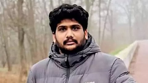 A medium shot of a young man with dark, wavy hair, a mustache, and a beard, standing outdoors in a misty or foggy wooded area. He is wearing a heavy grey puffer jacket with the brand text "CLOSURE LDN" visible on the left chest. The background features a blurred road and leafless trees, suggesting a cold or wintry climate.