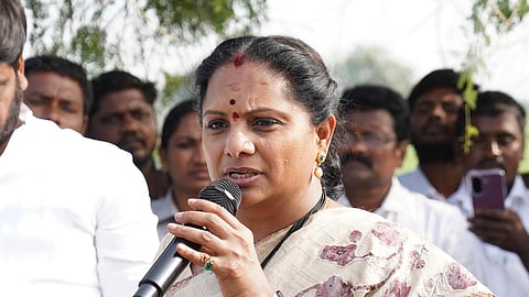A medium shot of Kalvakuntla Kavitha, an Indian politician, speaking into a black handheld microphone. She is wearing a cream-colored saree with maroon floral patterns and a gold border, paired with a black smartwatch and traditional gold jewelry. She has a serious expression and is gesturing with her right hand while addressing a crowd. Several people are visible in the blurred background, including a man in a white shirt and others watching her speak.