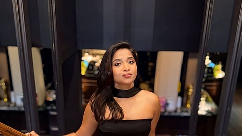 A young woman with long dark hair stands on a wooden staircase, wearing a sleek black strapless gown with a high-neck choker collar. She is looking directly at the camera with a confident expression, one hand resting on the wooden banister. The background is a dimly lit, sophisticated indoor setting with dark walls and subtle warm lighting.