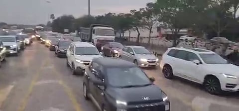 A blurry, low-angle, eye-level medium shot shows several lanes of vehicles, including a white truck, moving toward the camera on a multi-lane highway in Hyderabad, India. The vehicles are in a traffic jam on the Hyderabad-Vijayawada highway during the Sankranti festival. The sky is overcast. Trees and buildings line the right side of the road. Most of the vehicles are white, silver, or black. The lighting is dim.