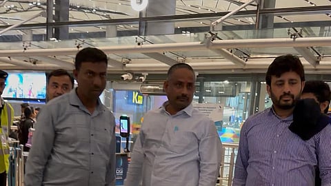 Three men stand in the middle of a brightly lit, modern indoor space that appears to be an airport terminal. The man on the left wears a light blue long-sleeved shirt and khaki pants. The man in the center wears a light-colored button-down shirt and grey trousers. The man on the right wears a blue and white striped long-sleeved shirt and dark grey pants. In the background, there are signs, a gate numbered "8," and other people moving through the terminal.