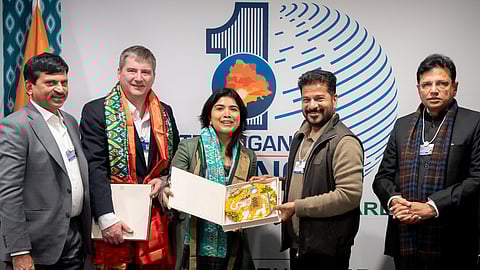 A group of five people stand in front of a white backdrop with a large blue logo and text. In the center, a woman in a green jacket and a man in a brown vest hold an open box containing a decorative plate featuring an elephant design. To the left, a man in a grey suit and another man wearing a colorful patterned shawl over a dark suit stand. To the right, a man in a black jacket and a blue and brown scarf stands with his hands clasped. The backdrop includes the names "Sri A. Revanth Reddy" and "Sri D. Sridhar Babu." A blue notebook and a black wallet are on a white table in the foreground.
