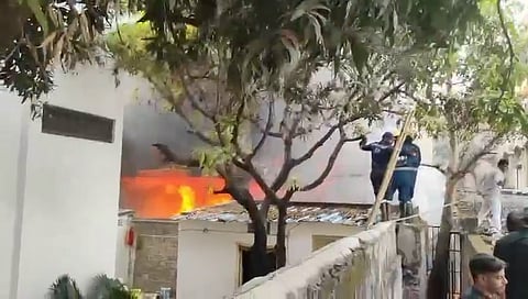 A medium shot shows a large fire burning in a building in Nampally, Hyderabad, India, on January 24, 2026. The fire is visible through a large window on the second floor of the building. The flames are bright orange and yellow, and thick black smoke is billowing out of the window. Two firefighters in dark blue uniforms and yellow helmets are standing on the roof of a smaller building in front of the burning building, directing a stream of water from a fire hose at the flames. A man in a white shirt and pants is standing on the ground to the right of the building, and another man is standing in the foreground, looking at the fire. A tree with green leaves is in the foreground, partially obscuring the view of the burning building. The building is made of white concrete and has a flat roof. The smaller building in front of it is also made of white concrete and has a corrugated metal roof. The sky is overcast and grey.