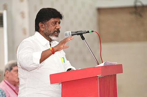 A man with a salt-and-pepper beard, wearing a white button-down shirt, stands behind a red podium and speaks into a microphone. He has his right hand raised in a gesturing motion. In the background, another man is partially visible, sitting and looking towards the speaker.