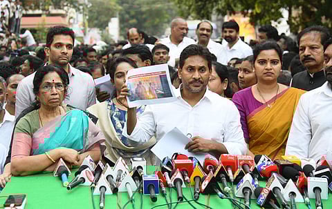 A high-angle, eye-level medium shot shows Y.S. Jagan Mohan Reddy, the former Chief Minister of Andhra Pradesh and leader of the YSR Congress Party, at a press conference. He is standing behind a table covered with a green cloth and crowded with numerous news microphones. He is wearing a white collared shirt and holding a piece of paper in his left hand.
To his right, a woman in a blue and white patterned top holds up a printed photograph for the cameras. To his left, another woman in a purple and orange saree looks toward the camera. An older woman in a green and pink saree is seated to the far left. Several other men and a dense crowd of people are visible in the background, many of whom are looking towards the center of the frame. The lighting is bright and even, suggesting an outdoor daytime setting.