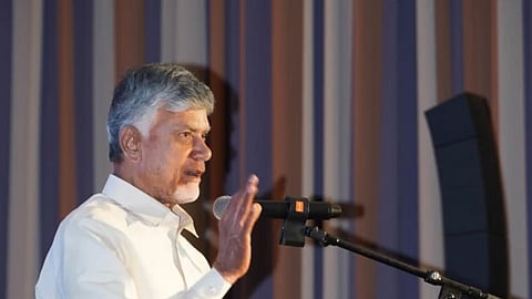 Chandrababu Naidu with grey hair and a light beard, wearing a white collared shirt, stands behind a podium and speaks into a microphone. He is gesturing with his right hand while holding a piece of paper in his left. The podium has a logo and the text "ANDHRA PRADESH India's Sunrise State" on it. The background consists of vertical brown and grey curtains.