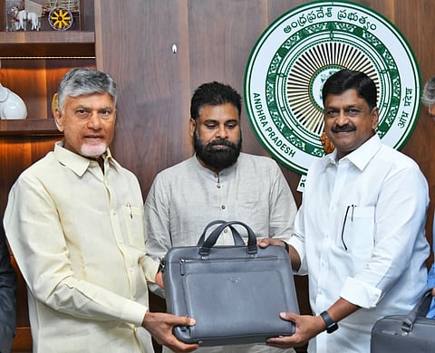 A medium shot of three prominent Indian politicians from Andhra Pradesh standing indoors against a wooden wall featuring the state government emblem. On the left, N. Chandrababu Naidu, in a light yellow shirt, and on the right, Payyavula Keshav, in a white shirt, are jointly holding a large grey leather briefcase (typically used for budget presentations). In the center, Pawan Kalyan, sporting a thick beard and a light grey kurta, looks on. The atmosphere is formal and significant, marking a key legislative or administrative event.