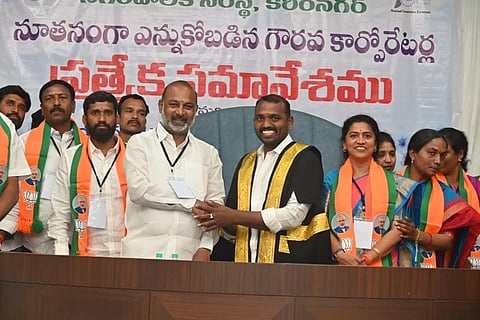 An image showing Bandi Sanjay Kumar, a prominent Indian politician from the Bharatiya Janata Party (BJP), shaking hands with a man dressed in a formal black and gold ceremonial robe, likely the newly elected Mayor or a high-ranking official of the Karimnagar Municipal Corporation. They are standing behind a wooden desk in front of a large banner in Telugu that mentions a "Special Meeting" for newly elected corporators. Several other people, including men and women wearing saffron and green scarves with the BJP lotus symbol and images of Prime Minister Narendra Modi, are standing behind them, smiling. The atmosphere appears to be one of celebration or an official swearing-in ceremony.