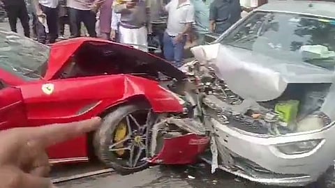 A red Ferrari with severe front-end damage is crashed into the front of a silver sedan on a city street. The Ferrari's front wheel is bent inward and its body panels are crumpled, while the sedan’s hood is crushed and its engine compartment exposed. Debris is scattered on the road, and a crowd of bystanders stands nearby observing the wreck. A hand in the foreground points toward the damaged Ferrari.