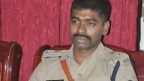 A medium shot shows Sunil Naik, an Indian Police Service (IPS) officer, from the waist up. He is seated in a dark wooden chair with a red cushioned back. He has a dark complexion, short black hair, and a thick black mustache. He is wearing a khaki police uniform with a nameplate on his left chest and a brown leather strap diagonally across his chest. He is looking off to the left with a serious expression. The background consists of a red curtain.