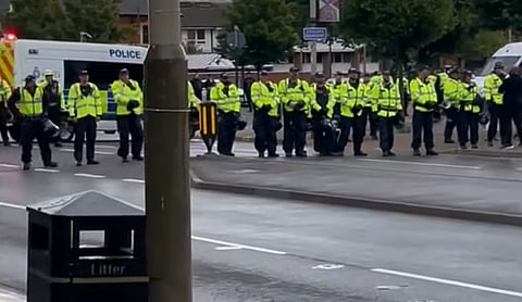 A wide shot shows a long line of police officers in high-visibility yellow jackets and dark trousers standing across a wet city street. They are positioned behind a dark grey litter bin and a utility pole in the foreground. In the background, a white police van with blue "POLICE" lettering is parked, and some trees and buildings are visible under an overcast sky.