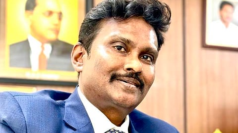 A medium shot of a middle-aged South Asian man with dark hair and a mustache, wearing a blue suit, white shirt, and a patterned blue tie. He is sitting in an office with his hands clasped, looking slightly away from the camera with a subtle smile. In the background, there is a framed portrait of B. R. Ambedkar on the wall and a small golden Buddha statue on a desk.