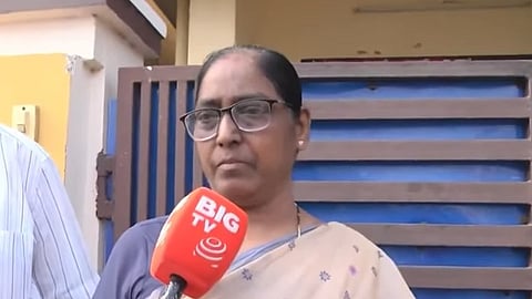 An eye-level, medium shot shows a woman with dark hair, glasses, and a light-colored saree being interviewed by a person holding an orange microphone with the BIG TV logo. She stands in front of a blue and brown slatted gate, and a person in a white striped shirt is partially visible on the left.