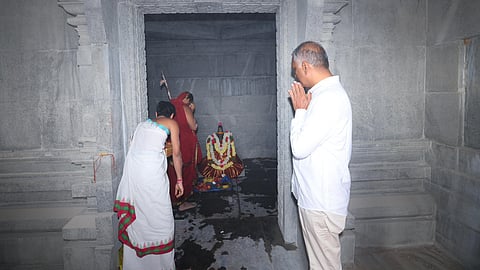 An indoor, eye-level shot shows T. Harish Rao, a man in a white long-sleeved shirt and light-colored pants, standing with his hands in prayer in front of a stone shrine. To his left, two people are inside the shrine, one wearing a white and red cloth and the other in a red cloth, facing a small, decorated deity. The shrine is made of grey stone with intricate carvings around the doorway. The floor inside the shrine is dark and wet.
