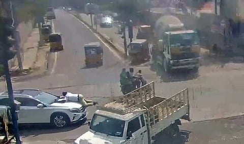 The scene depicts a chaotic moment of traffic:
In the center-left, a white light-duty truck with an open flatbed is angled across the lane.
To the left of the truck, a white SUV is stopped with a person appearing to lean over its hood.
In the center-right, a black motorcycle lies on its side in the middle of the road, suggesting a recent accident.
Various other vehicles are visible, including several yellow auto-rickshaws, a red hatchback, a dark car near the motorcycle, and a large green truck in the background.
The road surface is gray asphalt with faded white lane markings and a zebra crosswalk on the right. Buildings and a few trees line the far side of the intersection.