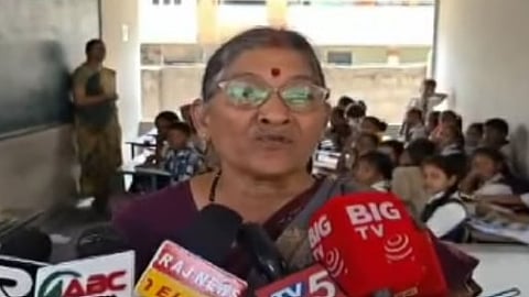 An elderly woman with gray hair, glasses, and a red bindi is speaking into several news microphones. She is in a classroom setting, with students visible sitting at desks in the background. Several news channel logos, including "BIG TV" and "RAJ NEWS," are visible on the microphones in the foreground.