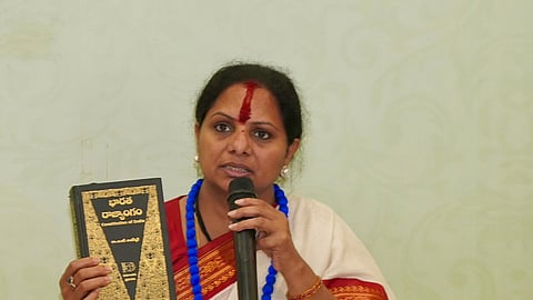 Kavitha Kalvakuntla with a red bindi and wearing a white and red saree is sitting down, holding a black book titled 'Constitution of India' in her right hand and a microphone in her left. There are several red and white signs in front of her with Telugu text. She is wearing a blue necklace and several gold bracelets. The background is a light green wall.