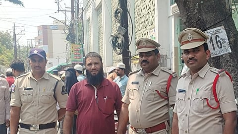 An outdoor, eye-level, medium shot shows four men standing on a city street. Three of the men are in tan police uniforms with matching caps and black or brown shoes. They stand side-by-side. On the far left, a police officer stands with his hands at his sides, looking directly at the camera. To his right, a man with a dark beard wearing a maroon polo shirt and blue jeans stands with his hands at his sides, looking at the camera. Next to him, another police officer in a tan uniform with a red shoulder cord stands with his hands at his sides, looking at the camera. On the far right, a third police officer in a tan uniform with a red shoulder cord stands with his hands at his sides, looking at the camera. In the foreground, a man in a white shirt is looking down at a cell phone in his hands. In the background, there are other people walking on the street, parked scooters, a large tree, and a building with a patterned wall. The lighting is bright and sunny.