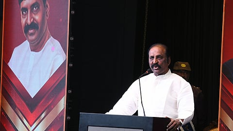 Vairamuthu with a dark mustache, wearing a white traditional kurta, stands behind a dark wooden podium speaking into a microphone. Behind him, to the left, is a large digital screen displaying a portrait of a younger man in a white shirt against a red and gold background. To the right, a security guard in a brown uniform is partially visible. The podium features a logo that includes the word "Leo."