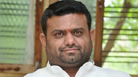 A medium shot of a man with short, dark hair and a groomed beard and mustache, wearing a white button-down shirt. He is looking directly at the camera with a neutral expression. The background is slightly blurred, showing what appears to be a window with wooden slats and some greenery outside.