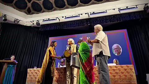 TNM journalist Nidhi Suresh, dressed in black with a mustard-yellow shawl, shakes hands with a presenter while receiving the Kamla Mankekar Award for Journalism on Gender 2025 on a stage in New Delhi. Other guests are seated and standing nearby, with the event backdrop visible behind them.