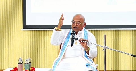 An elderly man with a bald head and glasses is seated behind a microphone, gesturing with his right hand raised while speaking. He is wearing a white traditional Indian outfit with a blue-and-white patterned shawl draped over his shoulders. Several water bottles are visible on a table to his left, and a large projection screen is in the background against a light-colored wood-paneled wall.