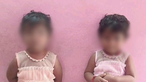 Two young children standing side-by-side against a pink wall, wearing matching ruffled sleeveless dresses. The child on the left wears a peach-colored dress, while the child on the right wears a light pink dress. Both dresses feature a sheer neckline with beaded detailing.