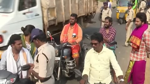 The image shows a busy street scene in Akividu, Andhra Pradesh, likely during a political or religious rally. Several men on motorcycles are being stopped or directed by police officers in khaki uniforms.
In the foreground, a man in a light yellow shirt is riding a motorcycle toward the camera, while behind him, another man wearing an orange shirt and a saffron scarf sits on a Royal Enfield motorcycle. To the right, a man is dressed in traditional attire, wearing a purple and gold dhoti with a matching shawl. A large white truck and a yellow auto-rickshaw are visible in the background, along with other pedestrians and police personnel managing the flow of traffic.