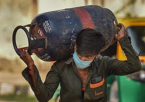A man wearing a green uniform shirt with an "Indane" logo and a light blue surgical mask carries a large, heavy blue and red commercial LPG cylinder on his right shoulder. He is using both hands to steady the cylinder, which has "19.0 kg" printed on its base. In the blurred background, a yellow and green auto-rickshaw is partially visible.