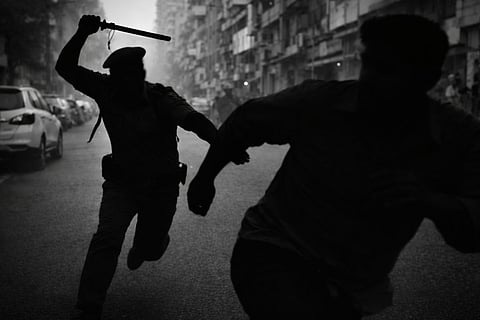 The silhouette of a man is seen with this arm raised to strike a blow. He is chasing another person who is fleeing from the man with the baton in hand. In the background is a street with buildings on either side, and cars parked on one side of the street.
