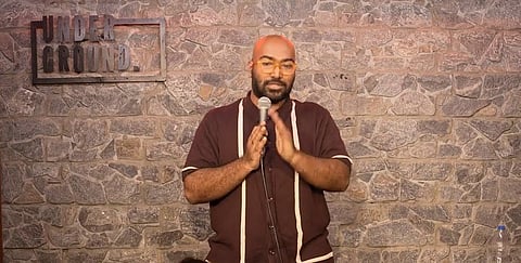 An image from a YouTube video featuring Indian stand-up comedian Anudeep Katikala. He is a bald man with a beard and glasses, wearing a maroon button-down shirt with vertical white stripes and beige trousers. He is standing on a stage with a stone-textured wall backdrop that has a sign reading "UNDERGROUND." He is holding a microphone with both hands and speaking to an audience, which is visible in silhouette in the foreground.