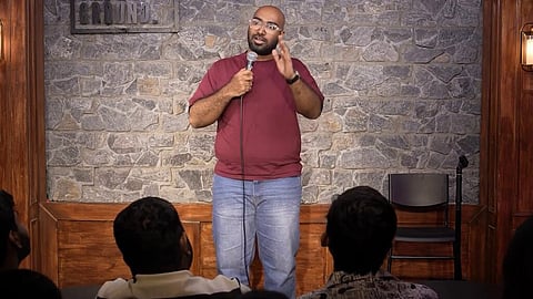 A stand-up comedian performing on a stage with a rustic stone wall backdrop. The man is bald, has a beard, and is wearing glasses, a maroon t-shirt, and blue jeans. He is holding a microphone and gesturing with his left hand while speaking to an audience, whose heads are visible in the foreground. A caption at the bottom of the frame reads "Migitha vaallu andharu," and a social media handle, @anudeepkatikala, is visible in the top right corner. Two warm sconce lights flank the stage area.