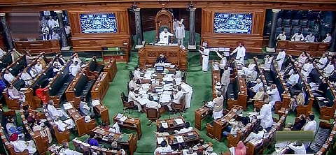 An overhead, wide-angle shot of the Lok Sabha chamber in India during a session. The room features ornate wood-paneled walls and a distinctive green carpeted floor.
At the far end, the Speaker sits in an elevated, carved wooden chair under a canopy, flanked by two large digital screens displaying a live feed of the proceedings. Below the Speaker, several officials are seated around a long, rectangular table cluttered with documents.
Members of Parliament are seated in curved, tiered rows of dark green benches on either side of the central aisle. Some members are standing and gesturing, while others are seated, reflecting an active legislative session. The lighting is bright and even, highlighting the architectural details of the chamber.