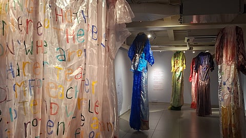 Large translucent garment with colourful handwritten text in the foreground, with multiple hanging textile installations behind in a gallery space.