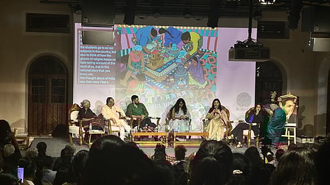 A panel discussion at the Building Begumpura conference in Bengaluru, with speakers seated on stage addressing an audience, as a screen behind them displays artwork and the conference title.