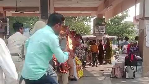 The image shows a busy bus station in India, with people gathered around a person who is on fire. The person on fire is standing in the middle of the crowd, and people are running away from them. There are also several police officers in the image, who are trying to put out the fire. The image is taken from a distance, and the quality is somewhat blurry. The background of the image shows several buses parked at the station. The overall mood of the image is one of chaos and panic.