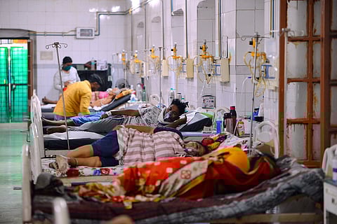 A wide shot of a crowded hospital ward with several patients lying in metal beds. Medical staff in yellow and white shirts attend to patients. Intravenous stands and oxygen equipment are visible along the tiled walls.
