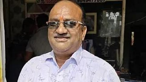 A man with a medium build and fair complexion is standing and looking directly at the camera with a slight smile. He has short, dark hair that is thinning on top, and he is wearing rectangular, rimless eyeglasses. He is dressed in a light purple, long-sleeved button-down shirt with a subtle floral or leafy pattern, featuring a cell phone and a pen tucked into the chest pocket. He also wears dark blue jeans, a black watch on his left wrist, and a black beaded bracelet on his right wrist. The background appears to be the entrance of a shop with various items and signs visible, though they are somewhat out of focus.