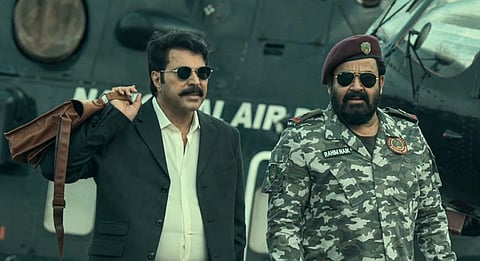 Actors Mammootty and Mohanlal stand side by side in front of a helicopter; Mammootty wears a black suit and sunglasses with a bag slung over his shoulder, while Mohanlal is in camouflage uniform with a maroon beret and sunglasses, indicating a military setting.