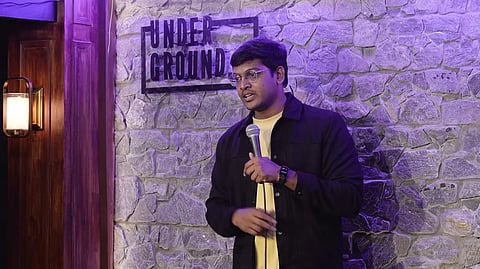 A man with glasses and a black jacket over a yellow shirt stands on a stage, holding a microphone. Behind him is a stone wall with a sign that says "UNDER GROUND". To the left is a wooden door with a light fixture and a painting of a woman. The scene is lit with purple light.