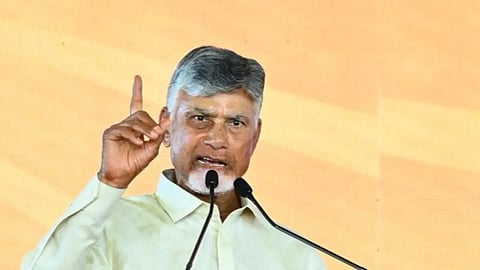 An image of N. Chandrababu Naidu, the leader of the Telugu Desam Party (TDP), addressing a crowd from a transparent podium. He is wearing a light yellow shirt, gesturing with his right index finger pointed upward, and speaking into two microphones. Behind him is a large backdrop featuring the TDP logo—a bicycle on a yellow field—and the party's name written in Telugu script on a red rectangular banner.