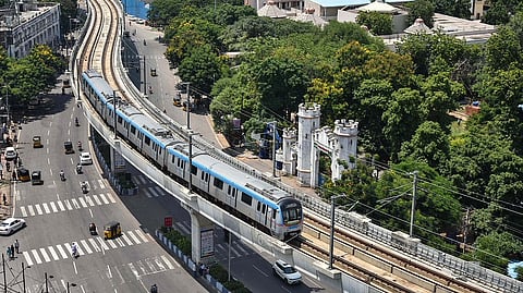 An aerial view shows a blue and silver Hyderabad Metro train traveling on an elevated track that curves through the city. Below the tracks is a multi-lane road with several white cars, yellow and black auto-rickshaws, and motorcycles. To the right of the tracks, there is a large area of lush green trees, behind which several buildings with traditional Indian architectural features and white domes are visible. A white, castle-like gate stands near the edge of the trees next to the road. The sky is bright, and the scene is captured in clear daylight.
