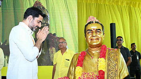 Nara Lokesh with a beard, wearing a white shirt, stands with his hands pressed together in a respectful namaste gesture before a golden bust of N.T. Rama Rao. The bust is adorned with red and yellow flower garlands. In the background, another man in a yellow shirt looks on, and security personnel stand behind the statue against a bright yellow backdrop.