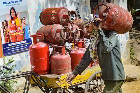 A person carrying an LPG gas cylinder. Behind him is a cart loaded with LPG cylinders
