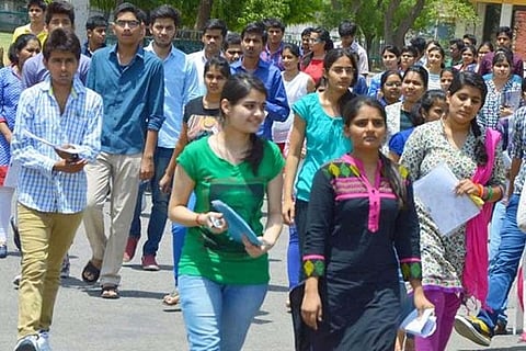 A large group of young Indian college students walks outdoors in a crowded, sunny scene. The students, both male and female, are dressed in a mix of casual and traditional Indian clothing.
Several female students are prominent in the foreground: one in a green top and blue jeans holding a blue folder, and another in a black kurta with pink and magenta embroidery holding papers. The male students are mostly in the background, with a few closer up wearing button-down shirts and t-shirts. They appear to be walking on a campus path. The image is a typical scene of students attending or leaving classes.