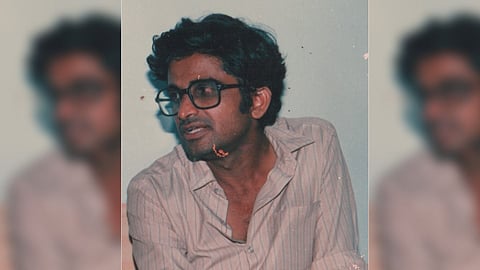 A medium close-up, slightly blurry, vintage-style photo of activist and intellectual K Balagopal. He is middle-aged, with dark, curly hair and is wearing thick-rimmed dark glasses. He is looking off to the left with a serious expression, and his mouth is slightly open as if speaking. He is wearing a light-colored, possibly beige or pale pink, striped collared shirt that is unbuttoned at the top. The background is a plain, light color.