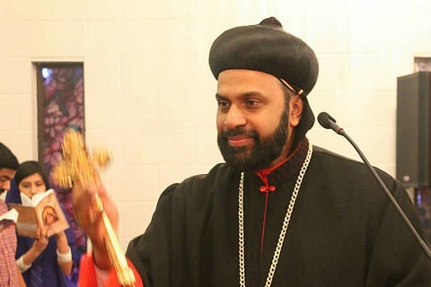 Bishop Kuriakose Mor Theophilos in black robes holding a golden cross in his right hand and standing near a microphone