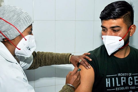 A health worker administers COVID-19 vaccine to a man in a black t-shirt. Both are wearing masks.