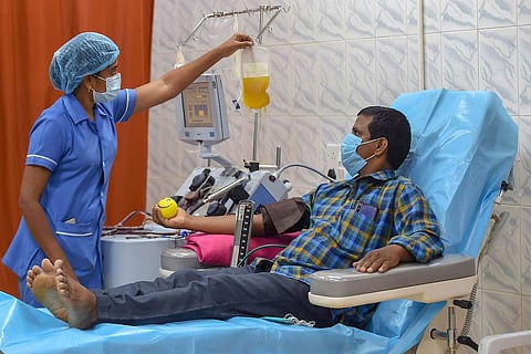 Man lying on hospital bed, nurse in blue next to him hanging a bag of plasm